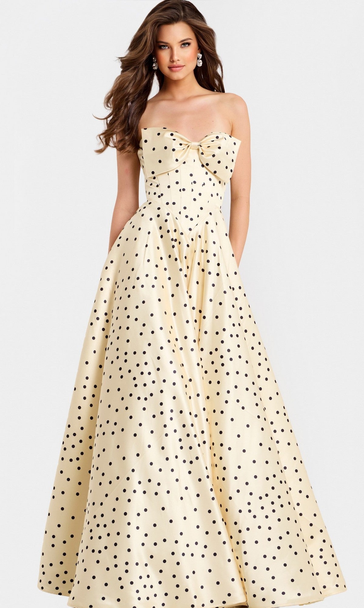 A woman stands against a plain background, glowing in the Jovani 49775—a champagne strapless satin ball gown with black polka dots and a charming bow detail on the bodice.
